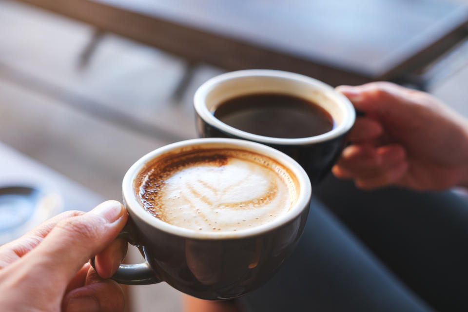 Two hands holding coffee cups, one has a latte with foam art and the other has black coffee