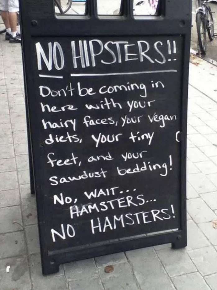 A sidewalk sign reads: "NO HIPSTERS!! Don't be coming in here with your hairy faces, your vegan diets, your tiny feet, and your sawdust bedding! No, wait... HAMSTERS... NO HAMSTERS!"