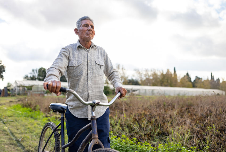 An elderly man in casual attire stands outdoors, holding a bicycle, with green fields and a greenhouse in the background