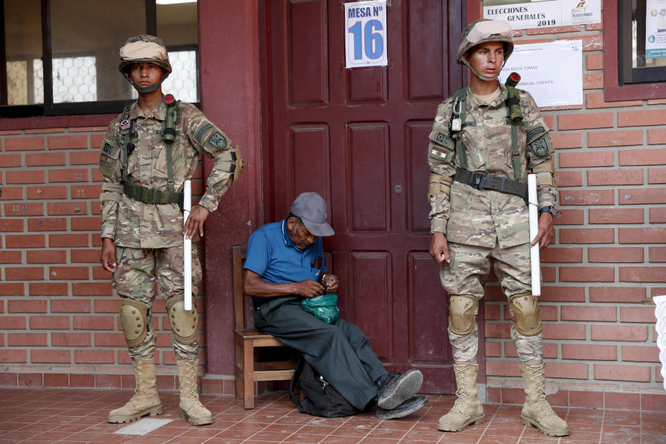 Military police guard the polling station where Bolivia's President Evo Morales will cast his vote as a man sits with a bag of coca leaves in Villa 14 de Septiembre, in the Chapare region, Bolivia, Sunday, Oct. 20, 2019. Bolivians are voting in general elections Sunday where Morales is Presidential candidate for a fourth term. (AP Photo/Juan Karita)