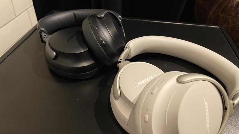 Two pairs of Bose QuietComfort Ultra Headphones (one black, one white) lying on a black surface by a white-tiled wall.