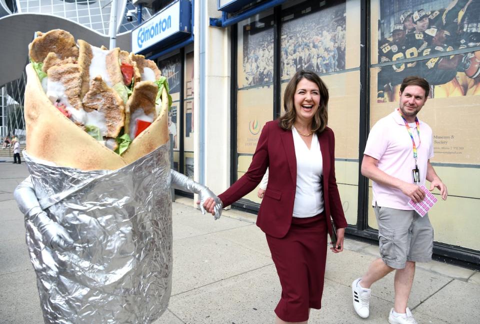 A premier walks with her snack muse — someone in a donair costume Alberta had once acquired for a distracted driving commercial, and this summer unloaded at a surplus auction.