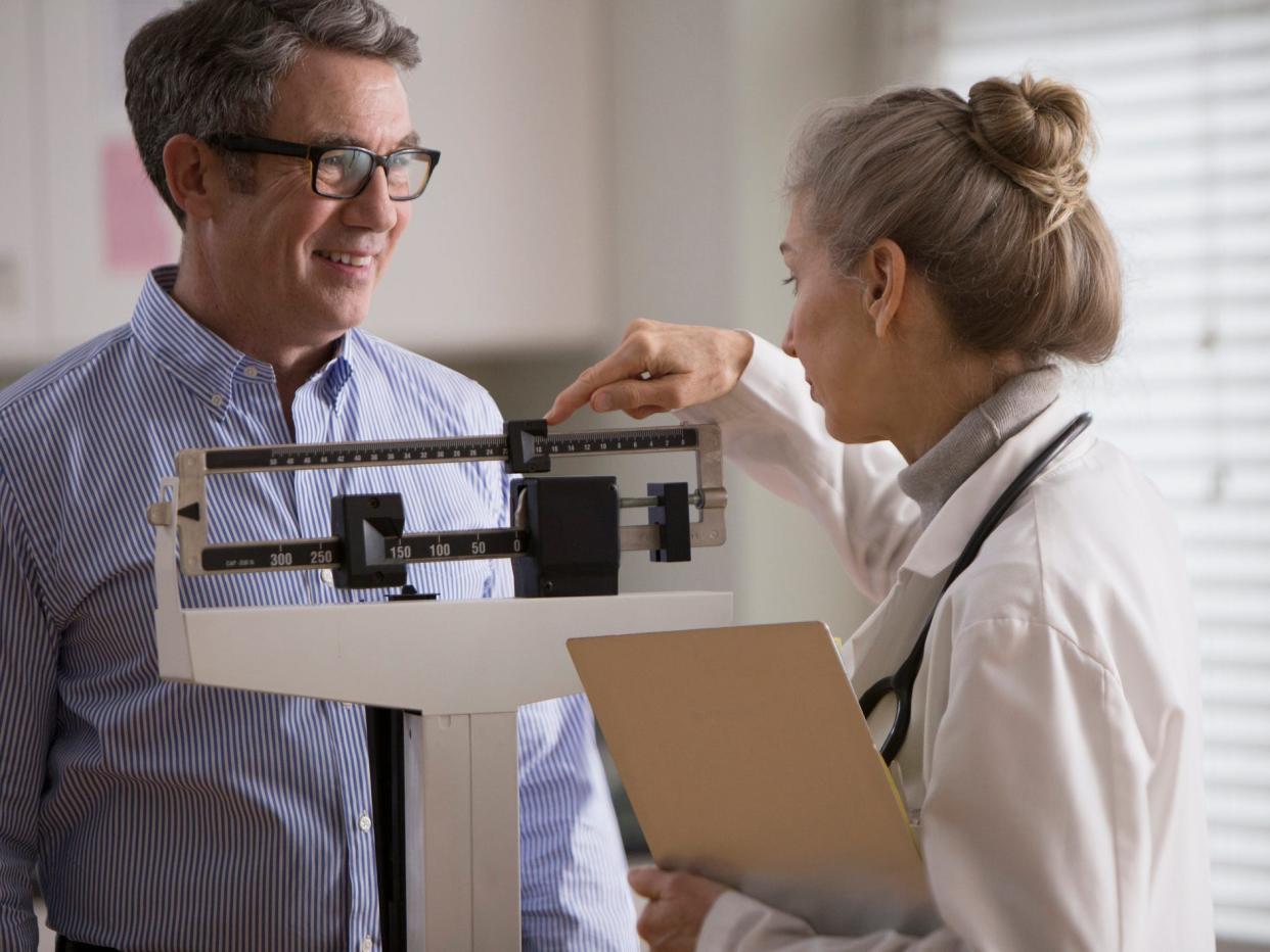 a older man at a doctor's office, standing on a scale, while a doctor takes weight loss measurements