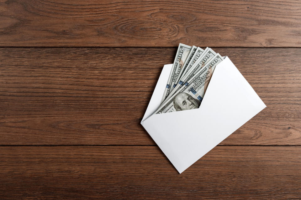 A white envelope partially open on a wooden surface, revealing several U.S. 100 dollar bills inside