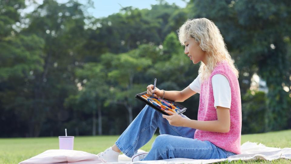 A woman sitting on a blanket in a park, using the LG Gram 2-in-1 tablet/laptop PC with a stylus.