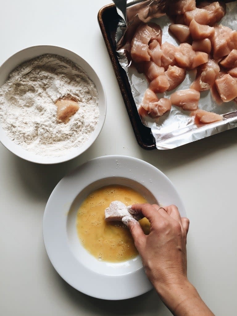 A person's hand breading chicken pieces in a bowl of egg wash next to a flour dish and tray of raw chicken