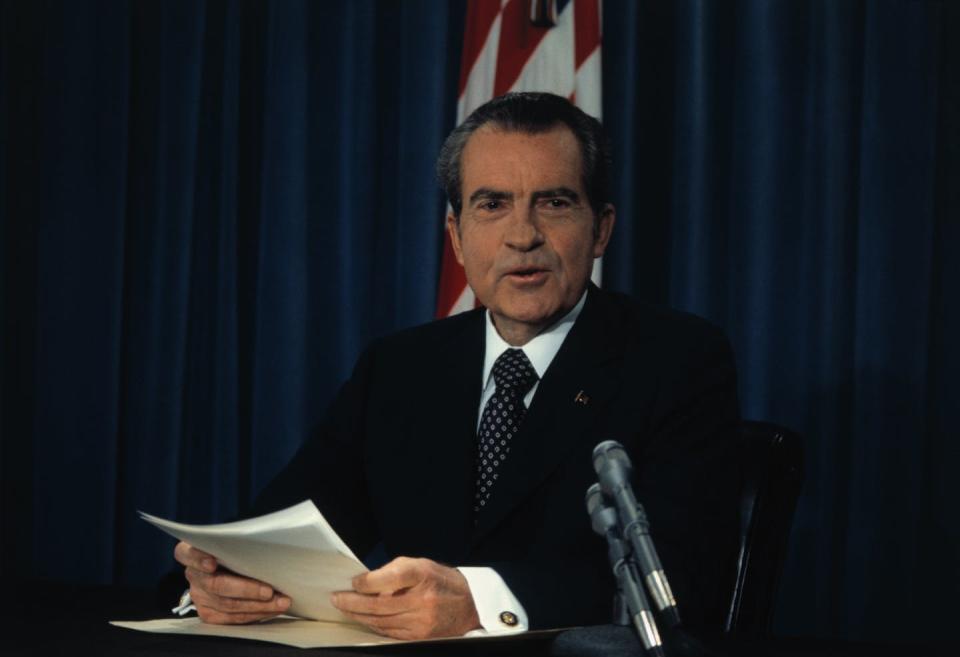 A man in a coat and tie sits at a desk holding papers.