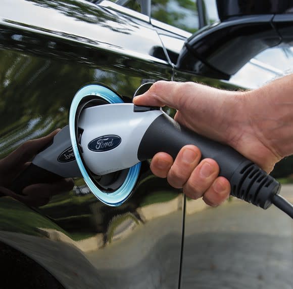 A Ford-branded electric charging plug is shown plugged into a Ford Fusion Energi plug-in hybrid sedan.