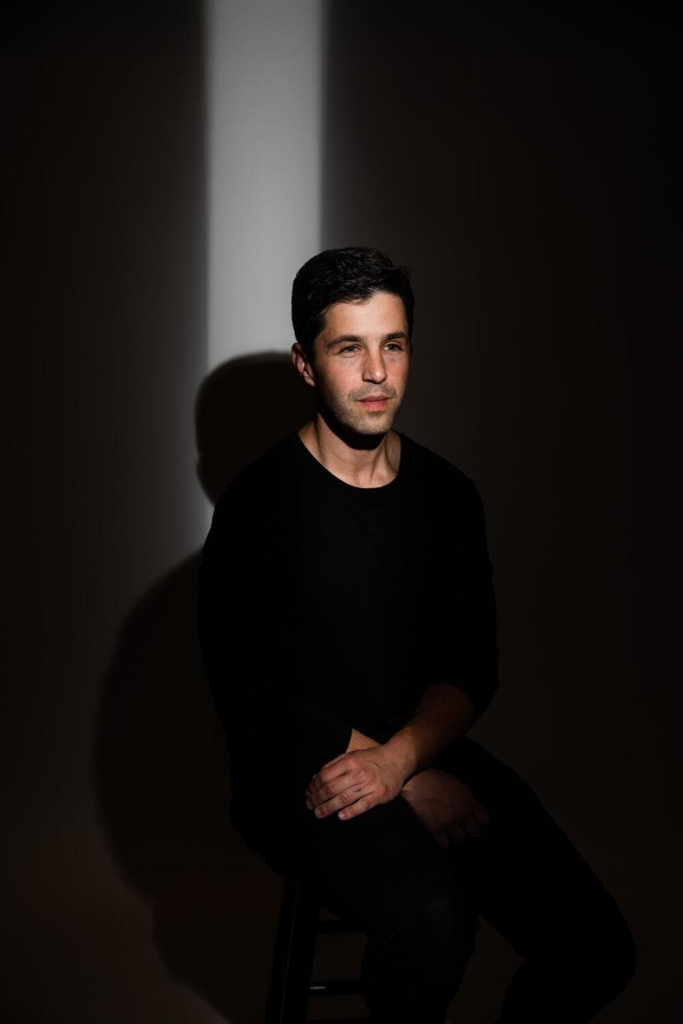 Josh Peck, in a black sweater, sits in a dark room.