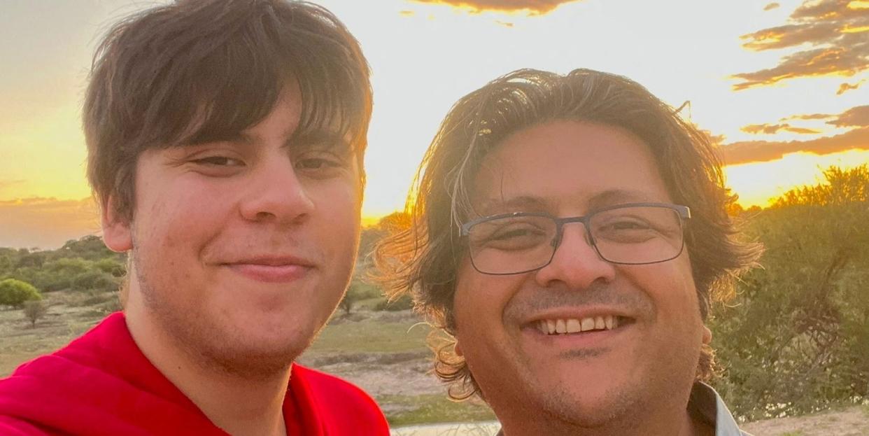 Suleman Dawood and his father, Shahzada Dawood, posing together on a sandy beach with some shrubs at sunset.