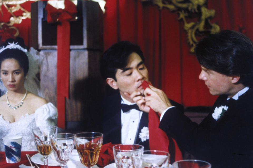 Actors in formal attire in a wedding scene from "The Wedding Banquet," directed by Ang Lee. The groom is being fed by another man while the bride looks on