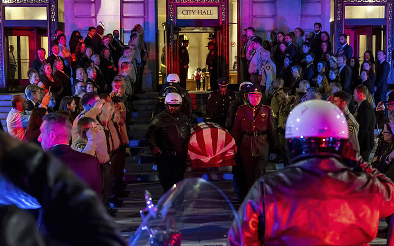 Honor guard members carry the casket of the late Sen. Dianne Feinstein (D-Calif.) down the steps of San Francisco City Hall. Members of law enforcement on either side salute, and the scene is lit by red and blue police lights.