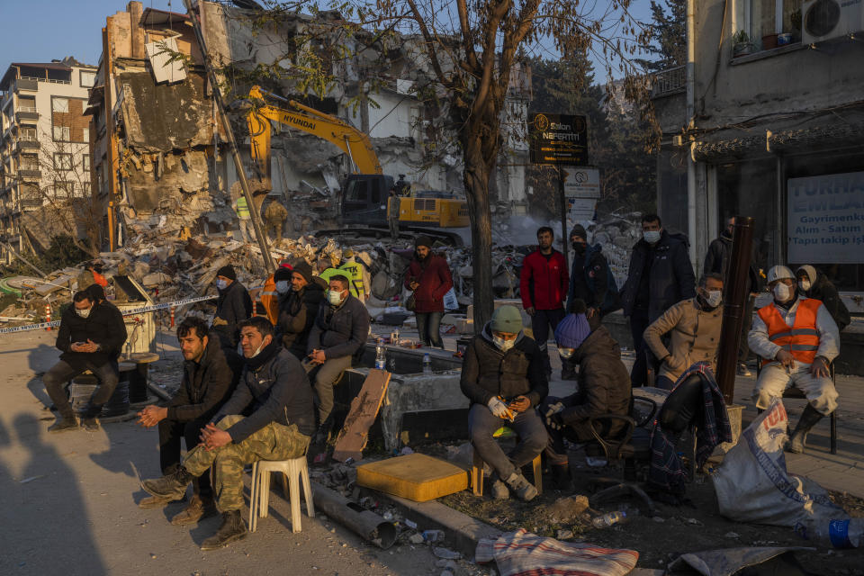 Varias personas sentadas el lunes 13 de febrero de 2023 junto a una vivienda destrozada mientras esperan que los cuerpos de sus amigos y familiares sean sacados de los escombros luego de un sismo en Antioquía, Turquía. (AP Foto/Bernat Armangue)