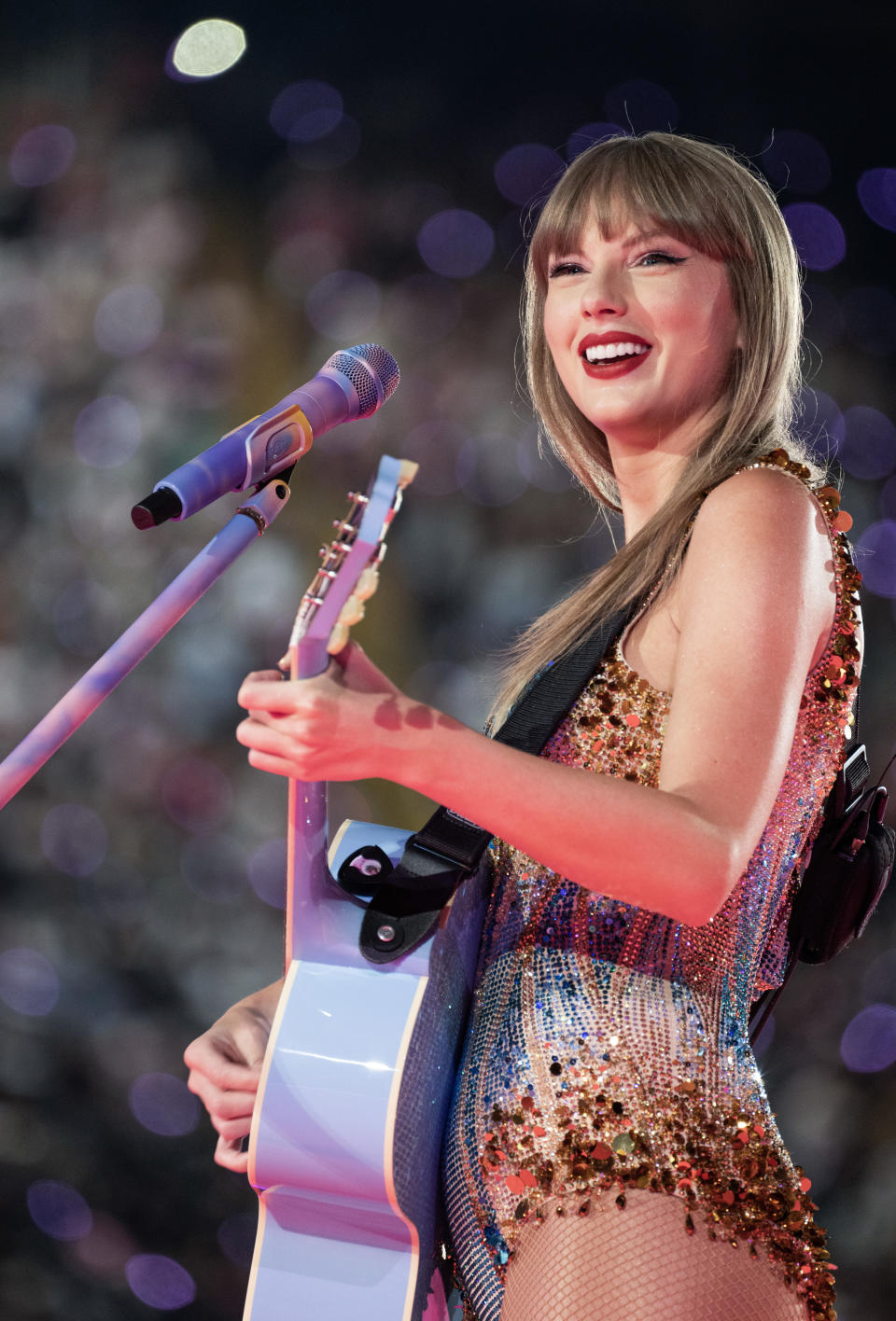 Taylor Swift performs on stage with a guitar and smiles at the audience. She wears a glittery, sequin-covered outfit