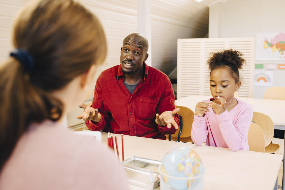 A man in a red shirt gestures while speaking to a woman in a pink top as a young girl focuses on a toy, in a classroom setting