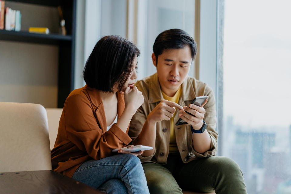 Image of Asian Chinese man and woman looking at smartphone and having a discussion in living room