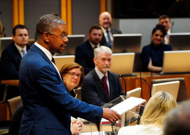 A man in a blue suit speaks to people sitting on benches 