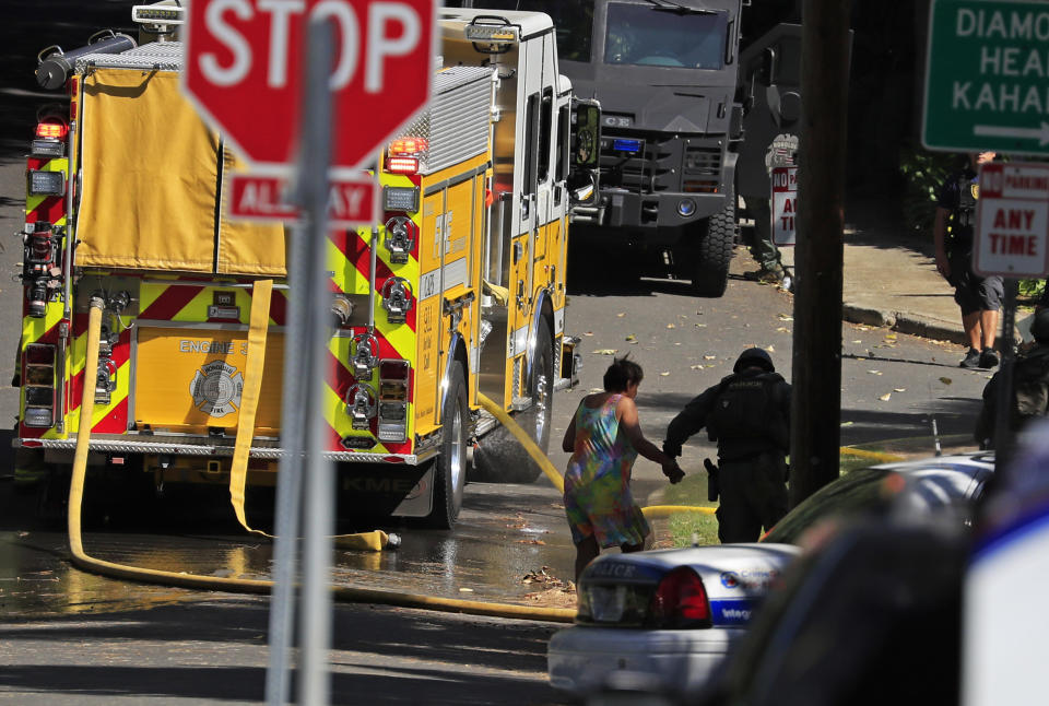 A Honolulu police officer escorts Dolores Sandvold back to her home near the scene of a deadly shooting on Sunday, Jan. 19, 2020, in Honolulu. Sandvold's home is located near the scene. A home a suspected gunman was believed to be inside caught fire and was quickly engulfed by flames. The fire at the home has since spread to several neighboring homes and a parked police vehicle. Sandvold said her house was not damaged. (AP Photo/Marco Garcia)