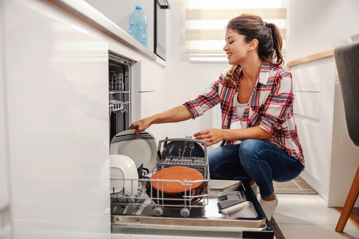 young woman squatting in kitchen and loading a dishwasher