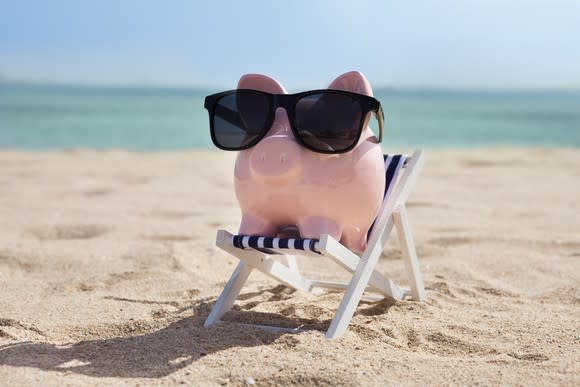 a piggy bank wears sunglasses on a little chair on a beach.