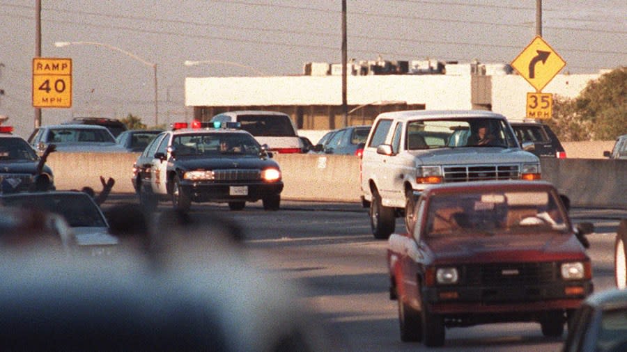 Motorists wave as police cars pursue the Ford Bronco (white, R) driven by Al Cowlings, carrying fugitive murder suspect O.J. Simpson, on a 90-minute slow-speed car chase June 17, 1994, on the 405 freeway in Los Angeles, California. Simpson’s friend Cowlings eventually drove Simpson home, with Simpson ducked under the back passenger seat, to Brentwood where he surrendered after a stand-off with police. (Credit: MIKE NELSON/AFP via Getty Images)