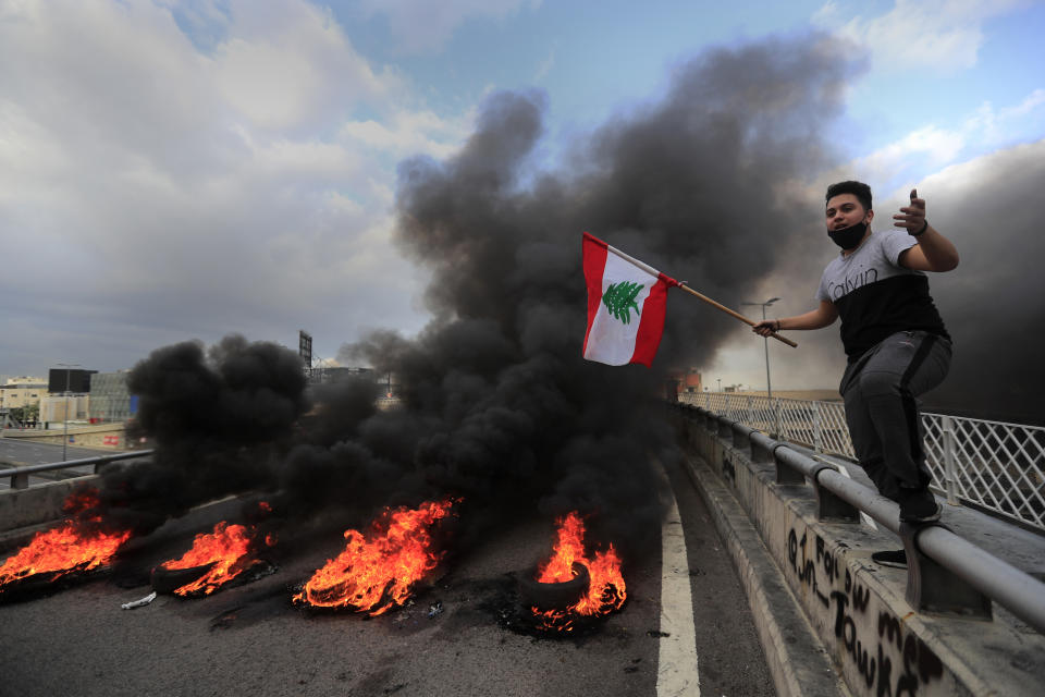 A protester waves a Lebanese flag near burning tires set to block a main highway, during a protest in the town of Jal el-Dib, north of Beirut, Lebanon, Monday, March 8, 2021. The dayslong protests intensified Monday amid a crash in the local currency, increase of consumer goods prices and political bickering between rival groups that has delayed the formation of a new government. (AP Photo/Hussein Malla)