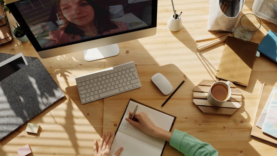 A woman's hand writing in a notebook on a desk while a woman's face appears on a computer screen for some online learning class thing.