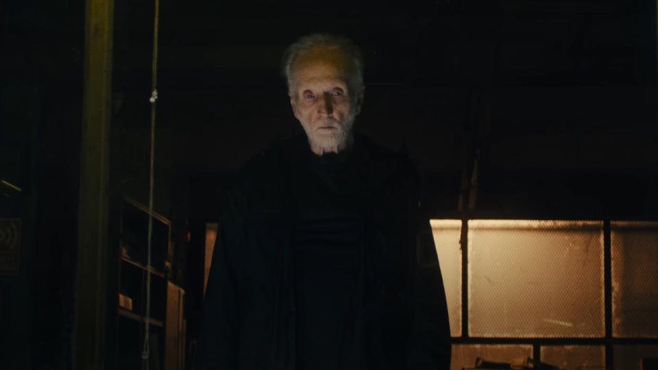 Tobin Bell stands ominously in a warehouse, dressed in black, in Saw X.