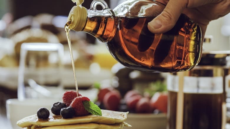 A hand pouring maple syrup out of a bottle onto pancakes