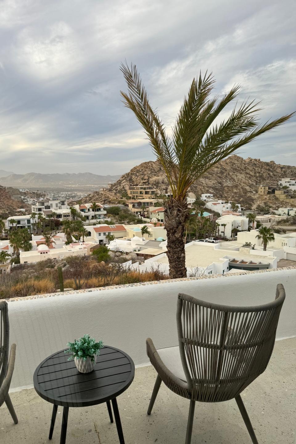 Two woven chairs and a small table with a potted plant overlook a scenic view of a hillside community featuring various houses and palm trees