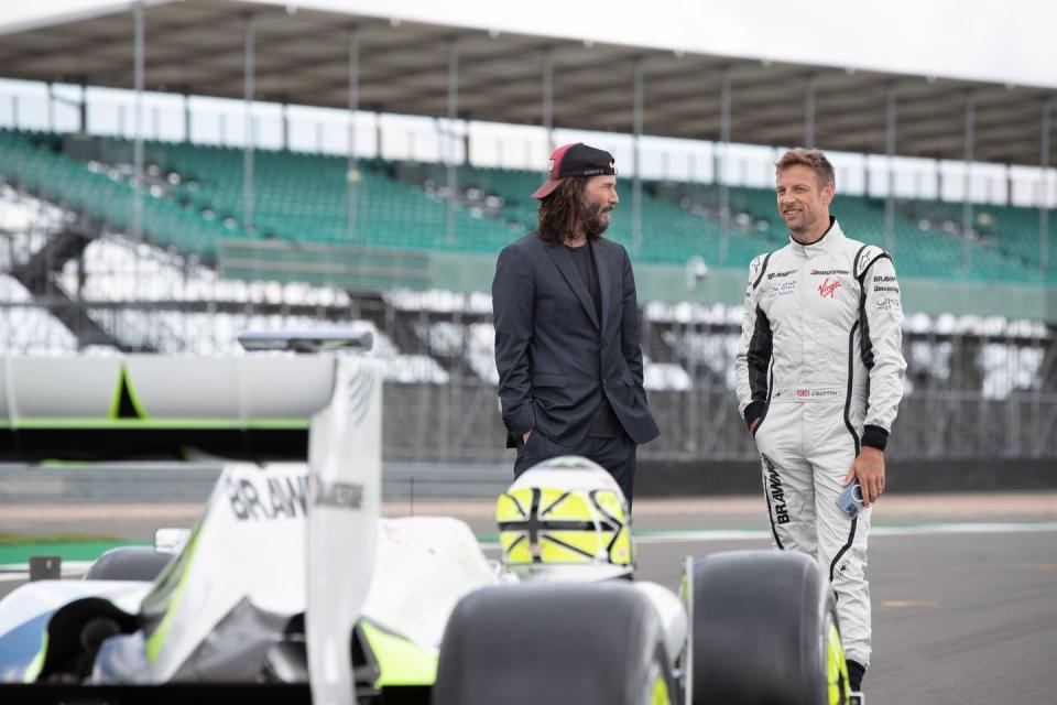 keanu reeves and jenson button talk to each other and smile as they stand behind a brawn formula 1 car on a racing circuit