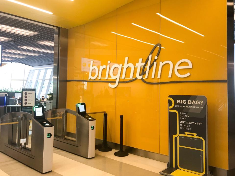 The Brightline logo against a yellow wall, with electronic ticket entrances and a sign about baggage limits. 