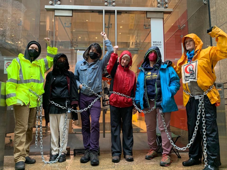 six people wearing jackets hoods stand in a line chained together at the hips in front of glass doors holding up their fists
