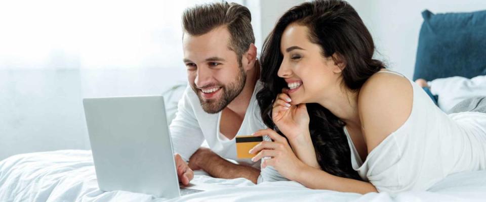 smiling young couple using laptop and holding credit card in bed, online shopping