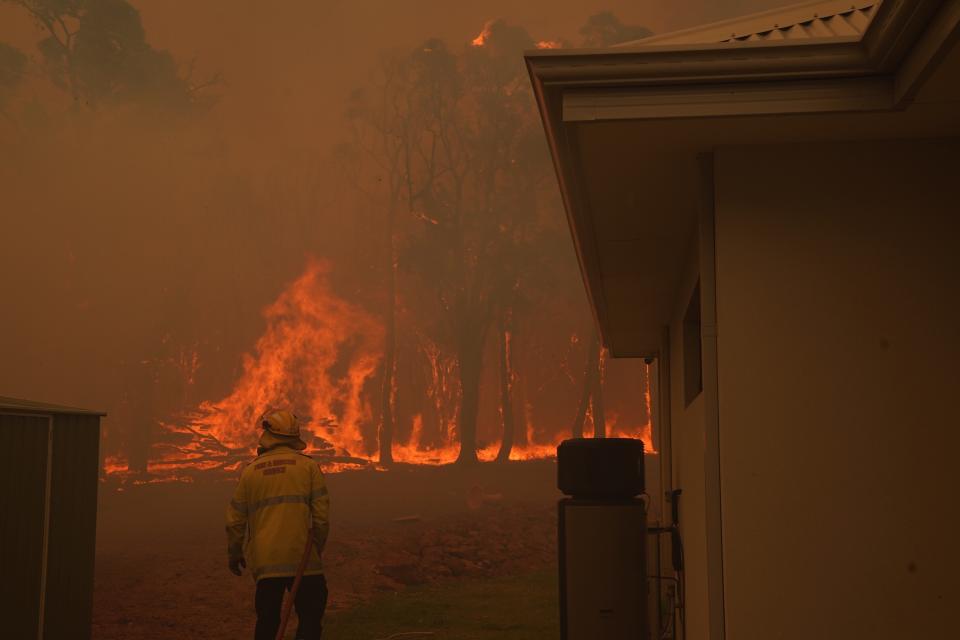 A firefighter standing next to a house. Fire burns in the background. 