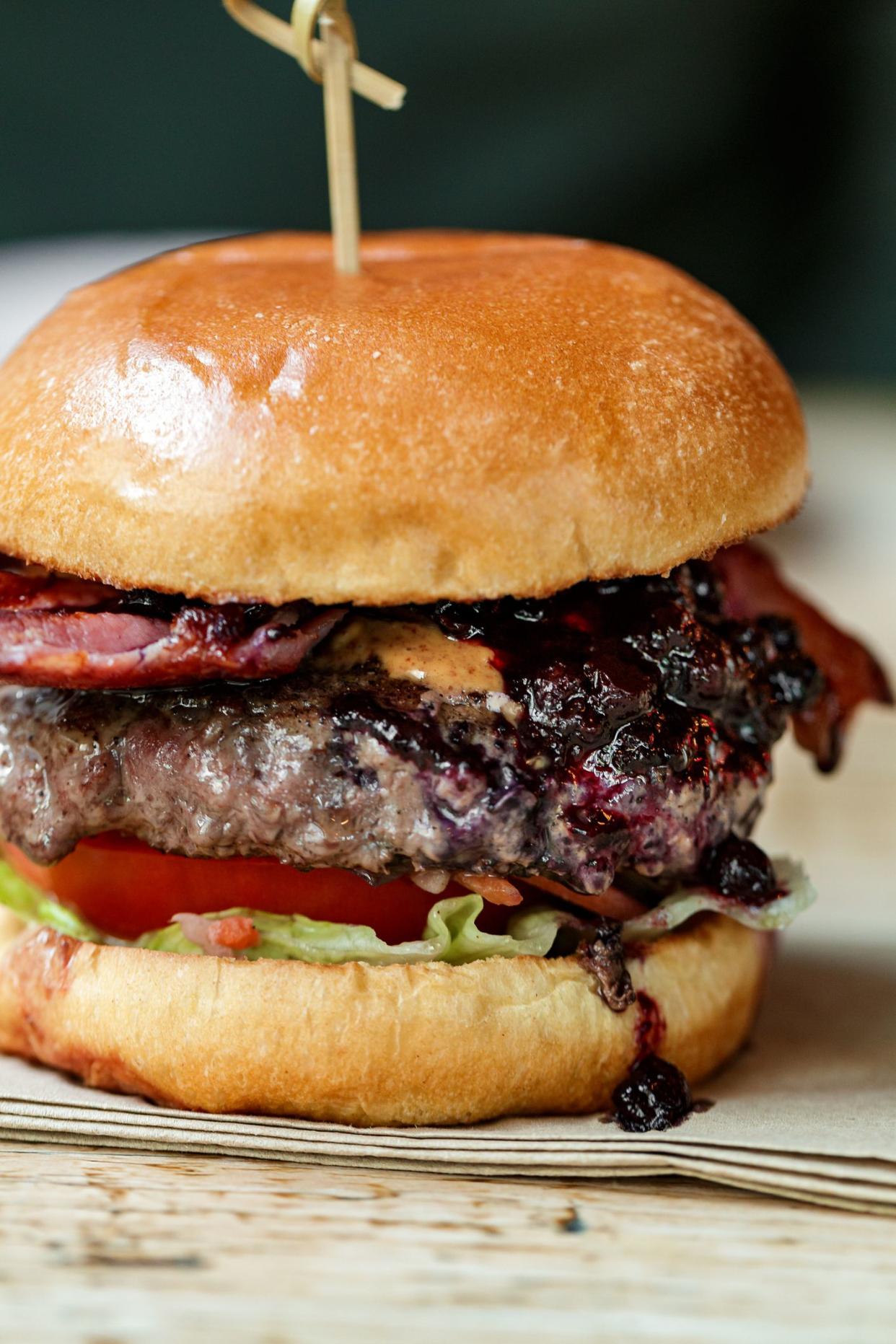 Close up image depicting a freshly cooked bacon cheeseburger in a restaurant. The burger is sandwiched between two glazed buns and is loaded with a flame grilled beef patty, melted cheese, blueberry jam and crispy bacon and salad.