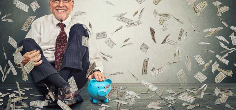 An older man sits by his piggy bank as money flies through the air.