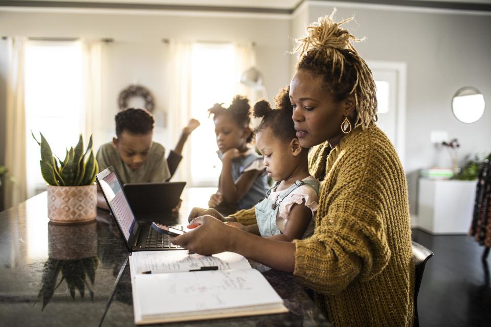 Mother working from home while holding toddler, family in background