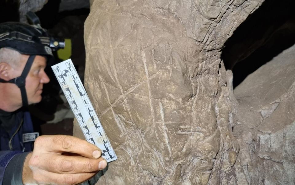 Explorer Lee Berger holds a photographic scale next to a crosshatched engraving in the passage between the Hill Antechamber burial chamber and the Dinaledi burial chamber in South Africa's Rising Star cave system - Mathabela Tsikoane/Guzelian