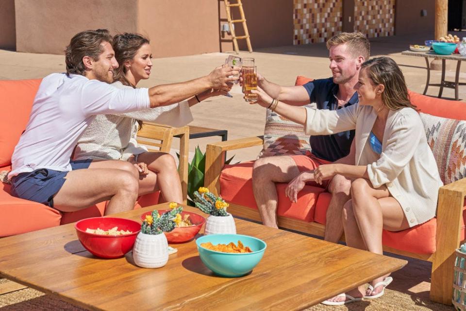 Four people toast with beer glasses sitting outside on a patio.