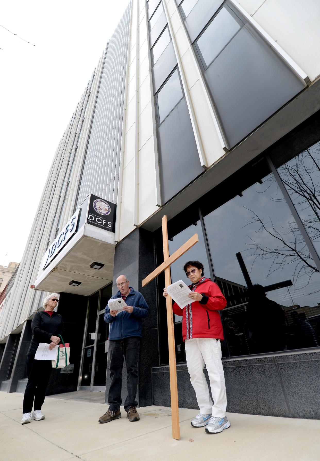 The fourth station of the cross is read by Chuck Weiss of Clinton, Mississippi, center, in front of the Department of Children and Family services as Vicky Bean of Springfield, right, holds a cross and Sister Marcelline Koch of Springfield watches during the 2022 Way of the Cross, which drew about 40 participants. Friday's walk starts at the Illinois Supreme Court building at Second and Capitol at noon.