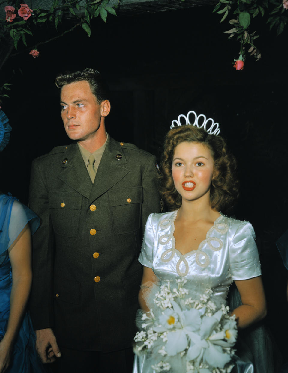 Man in military uniform beside a woman in a tiara and white dress holding flowers. They are both glancing to their left