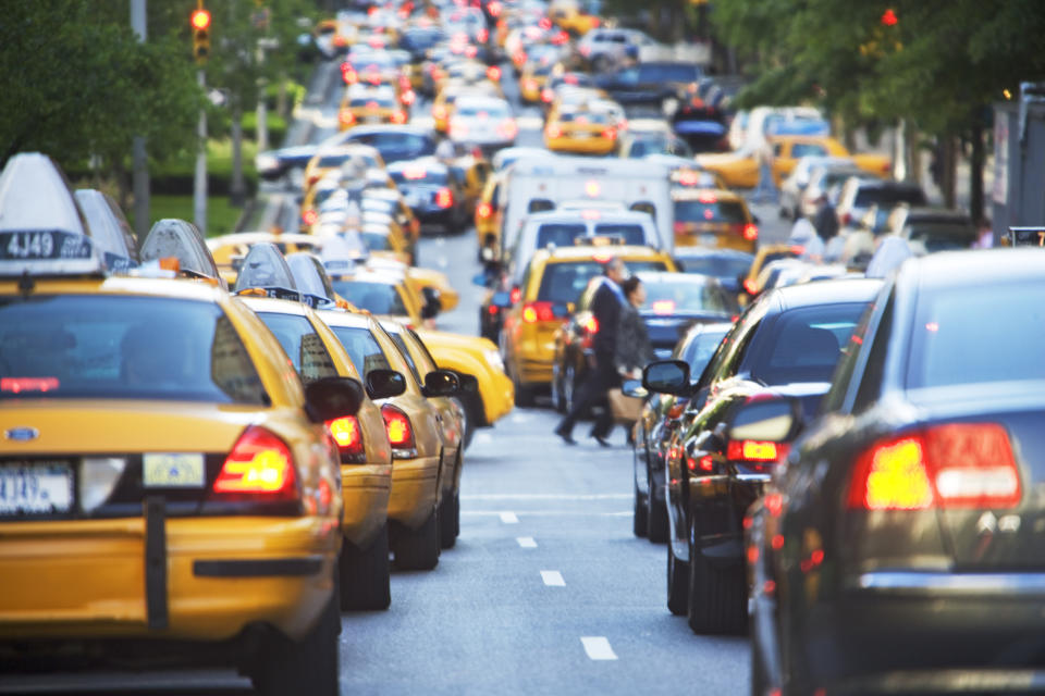 A busy city street full of yellow taxis and other cars. Pedestrians cross the road between the vehicles amidst the traffic jam.