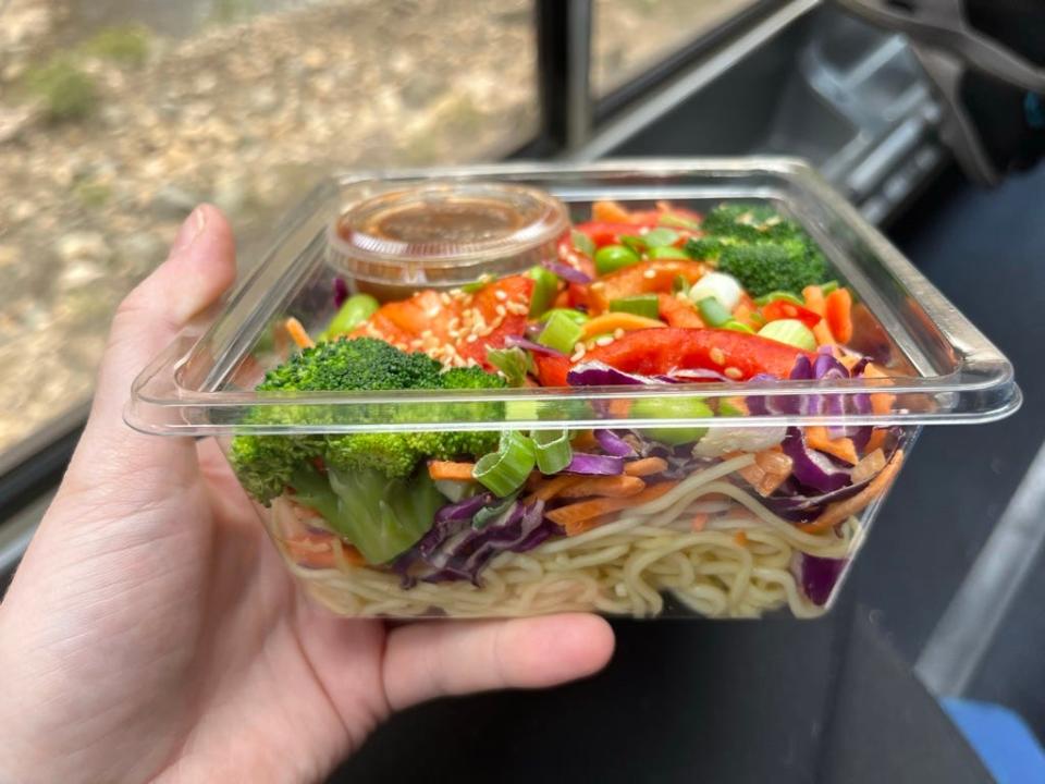 a veggie and noodle salad being held in front of a window on Amtrak
