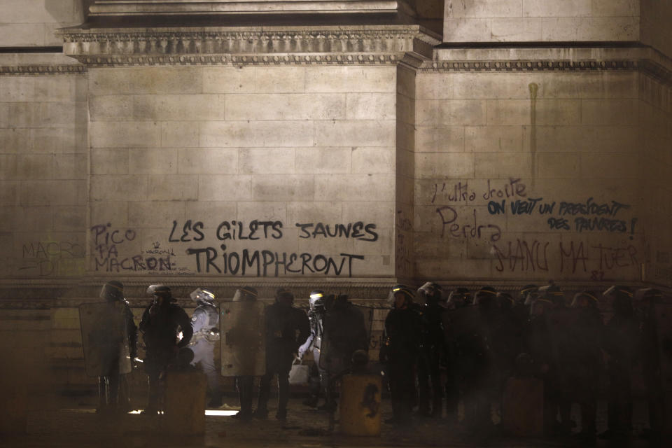 Riot police officers walk by the Arc de Triomphe tagges with the words "The yellow jackets will triumph", center, Saturday, Dec.1, 2018 in Paris. A French protest against rising taxes and the high cost of living turned into a riot Saturday in Paris as police fired tear gas and water cannon in street battles with activists wearing the fluorescent yellow vests of a new movement. (AP Photo/Kamil Zihnioglu)