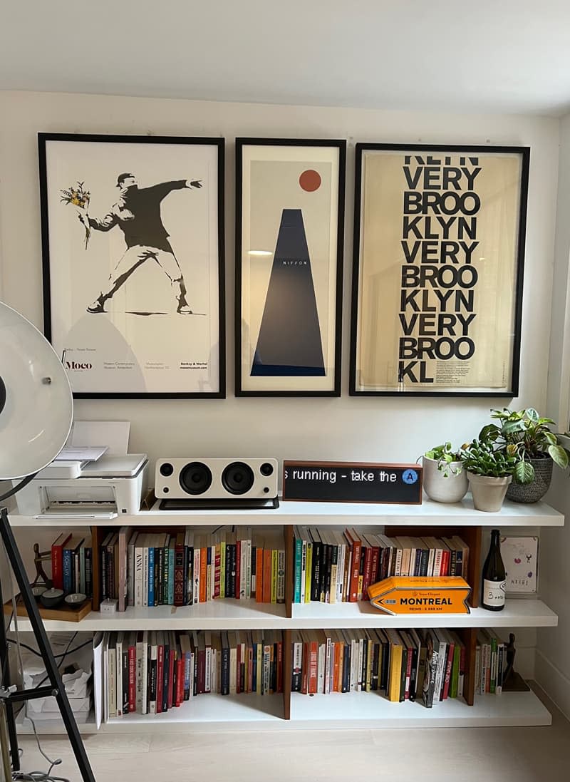 Art hung on neutral wall above bookcases topped with potted plants, speaker, and printer.