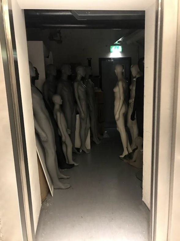 A dimly lit storage area containing numerous mannequins of various sizes positioned closely together, creating an eerie appearance