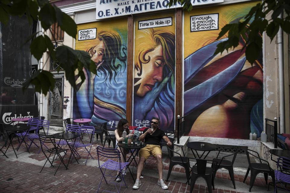 People sit at a cafe in front of a graffiti covering the fence of a closed old textile shop in Athens, Greece, on Sunday, June 7, 2015.  (AP Photo/Yorgos Karahalis)