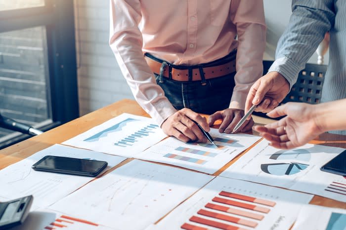 Pointing at financial charts, several people stand around a table.