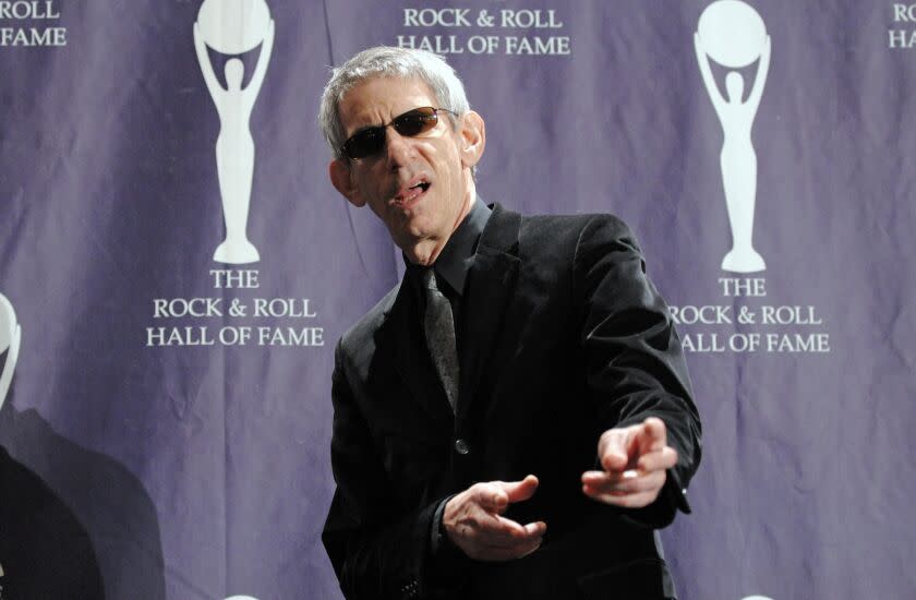 A man with gray hair wearing sunglasses and a black suit and pointing finger guns toward the camera